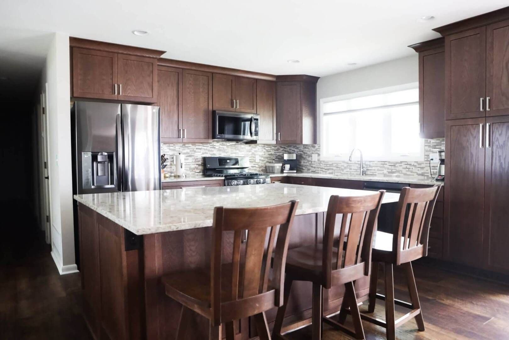 full image of dark wood kitchen cabinets, kitchen island, drawers and counter-height chairs with dark silver appliances.