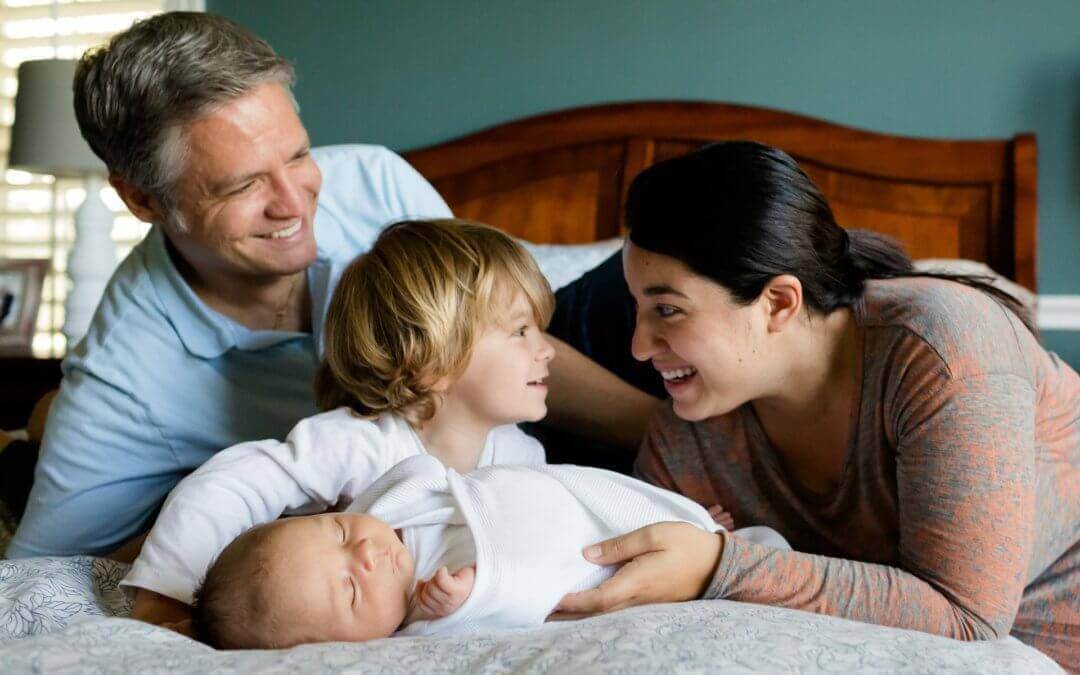 A man, woman, child, and infant snuggling, smiling, and laughing on a bed.