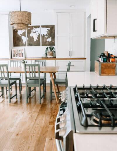 dark metal stovetop on white counter with white tile backsplash and small wooden box on counter for utensils. Wooden dining room table with 6 green chairs and a wood rattan pendant hanging above, white shelving along the back wall with wooden map wall art