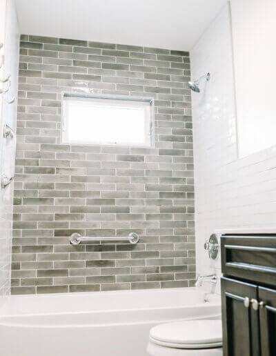 dark black wood countertop in right corner of image with white tiling along walls, gray accent tile wall at end of photo with white bathtub and toilet. 3 silver hooks along left wall for towels.