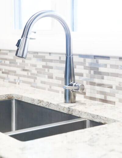 close up image of metal sink and silver faucet with multi-colored tile backsplash and white gray marble countertops